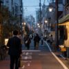 Local shoppers and commuters move through a neighborhood commercial street at dusk.