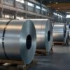 Unlabeled galvanized steel coils stacked on pallets in a quiet industrial warehouse with workers in the distance.