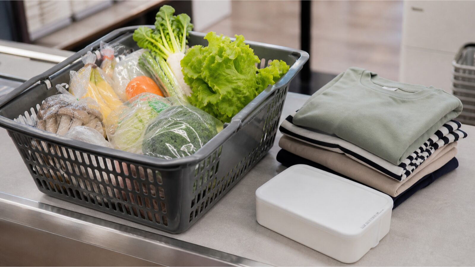Editorial retail still life with a shopping basket, fresh produce and folded clothing on a neutral checkout counter.
