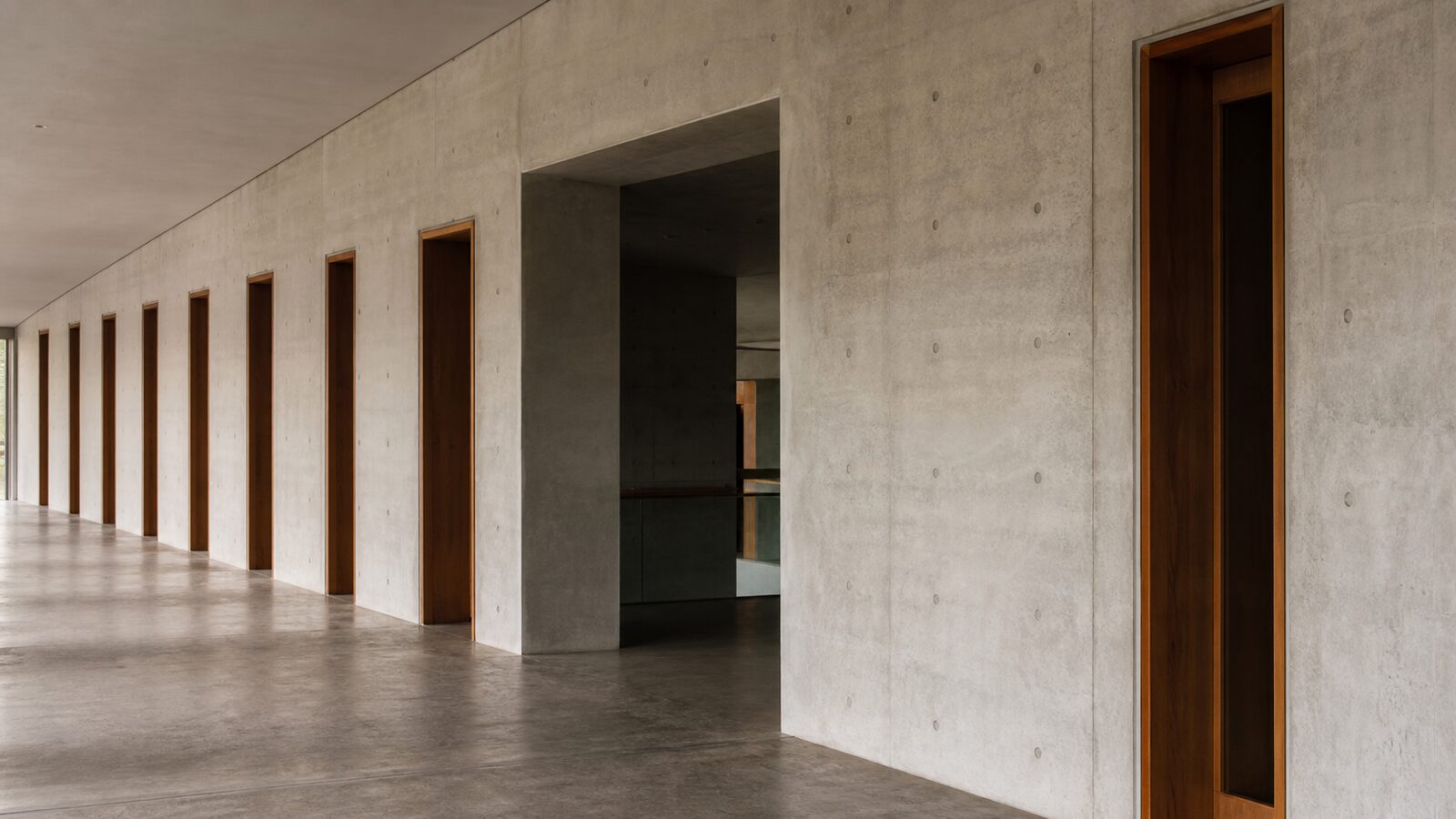 A concrete corridor has regularly spaced doorways and one visibly wider opening.
