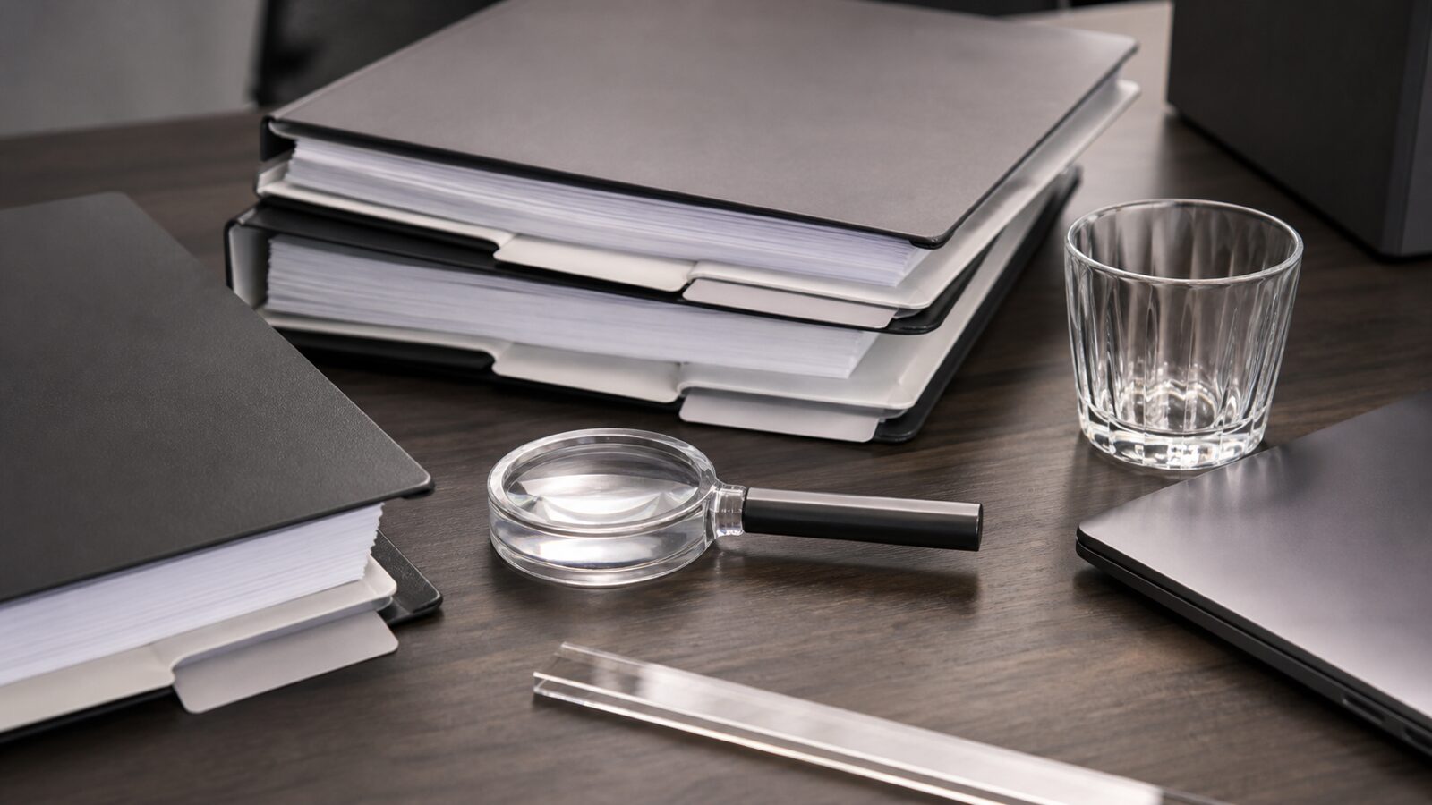 Compliance review desk with blank binders, a magnifying loupe, a clear ruler and a closed laptop.
