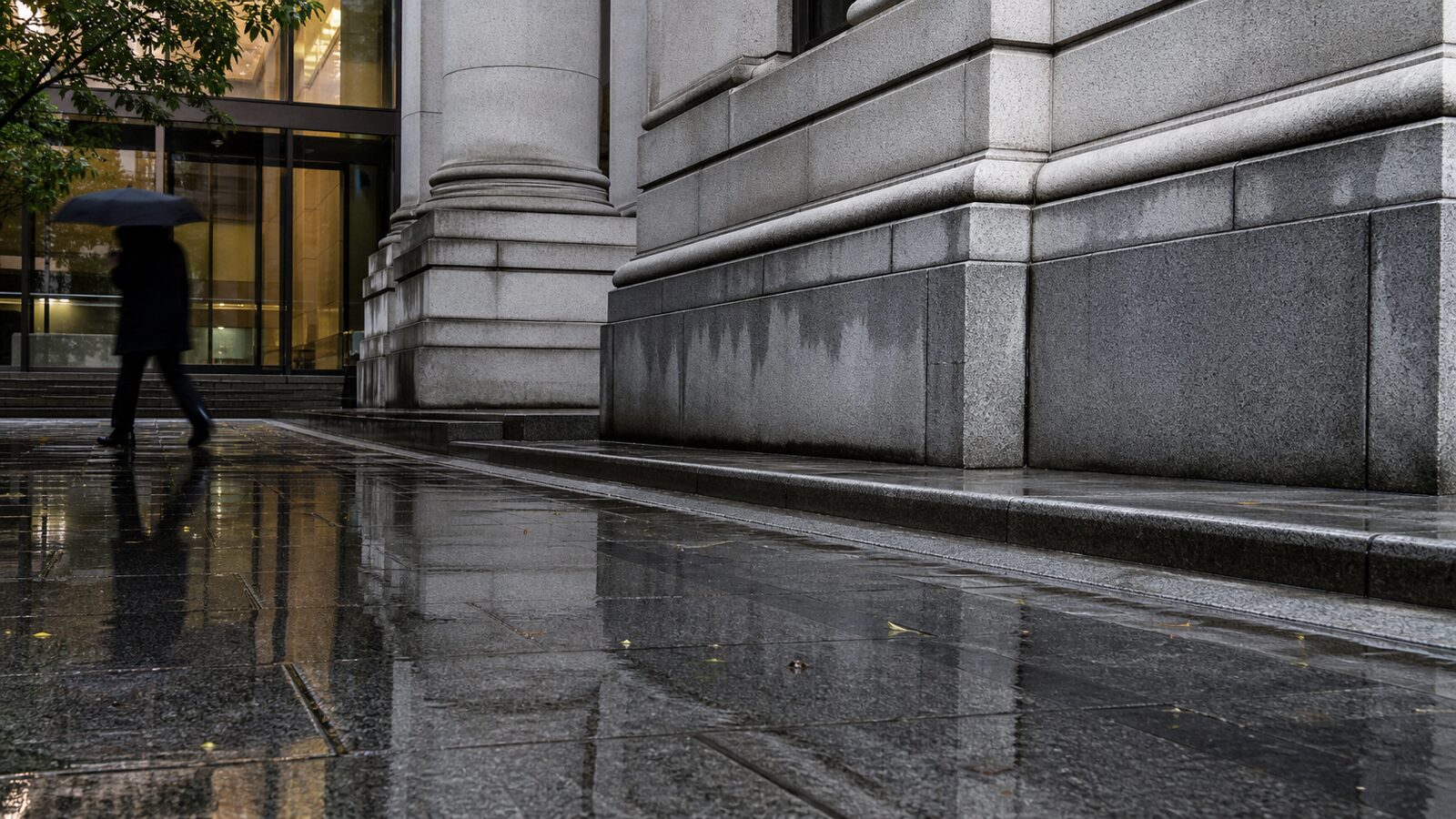 A rain-wet institutional stone building facade with a pedestrian silhouette