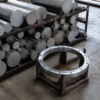 Matte aluminum billets on a worn steel rack beside a finished circular machined ring in a metalworking shop.