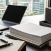 A quiet institutional investor desk with blank papers, a dark laptop screen, a calculator and folders near a city office window