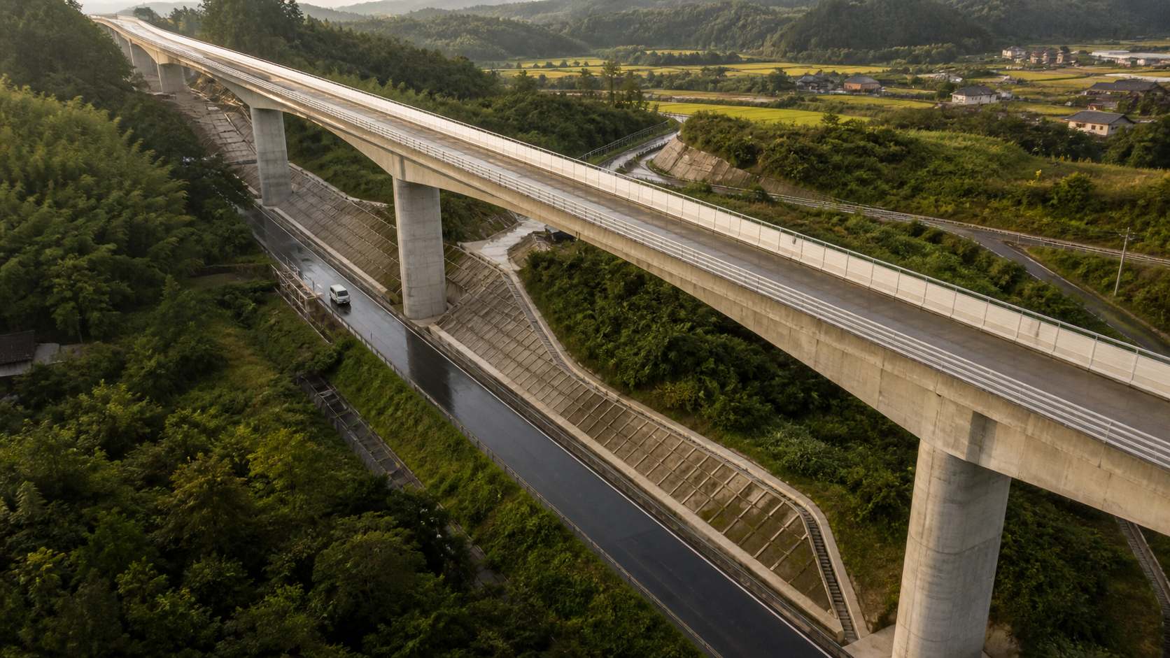 A modern concrete viaduct crosses above an older regional road and maintained retaining slopes after rain.