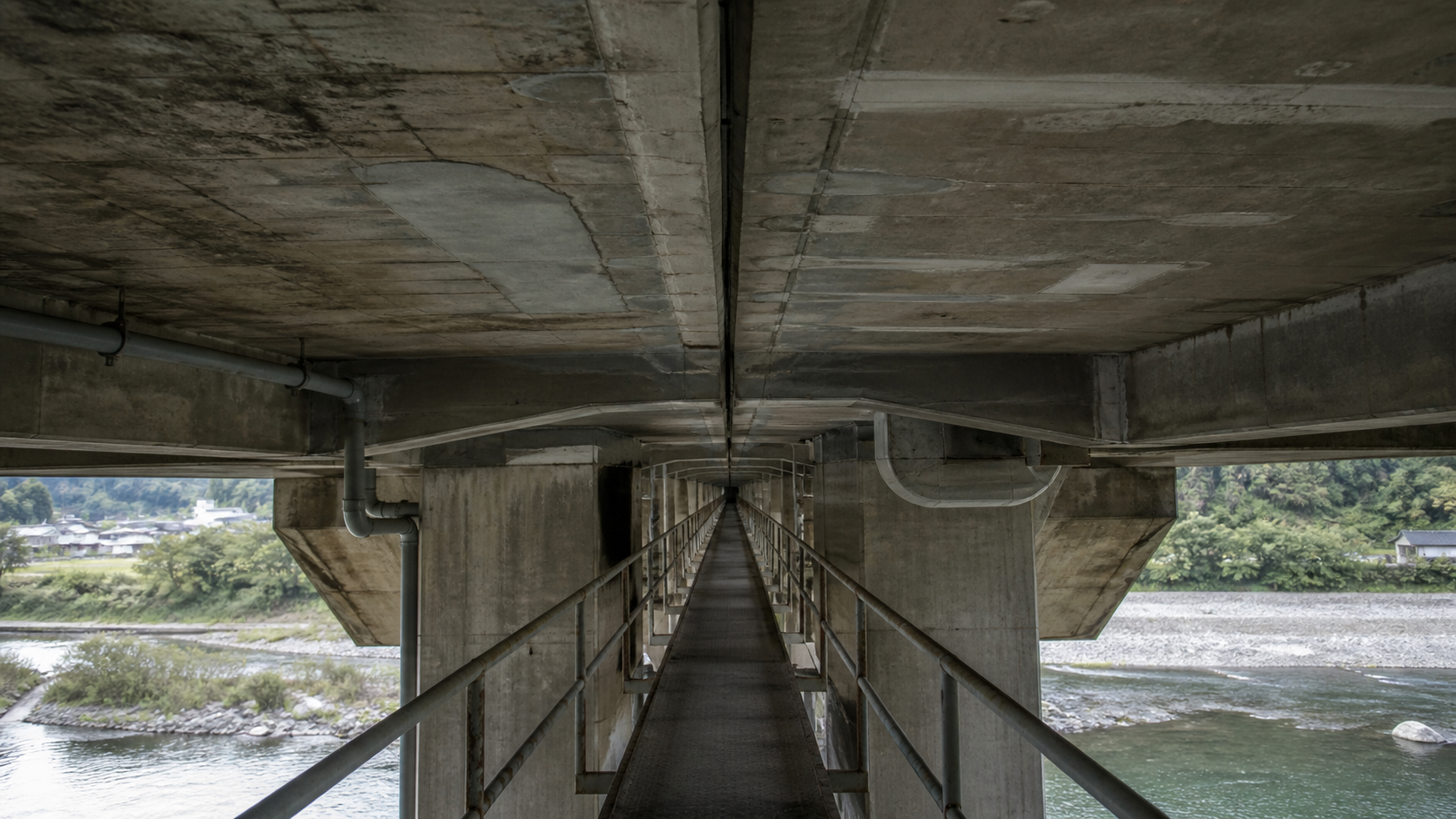 An empty inspection catwalk runs beneath a weathered concrete bridge with visible repairs and drainage pipes.