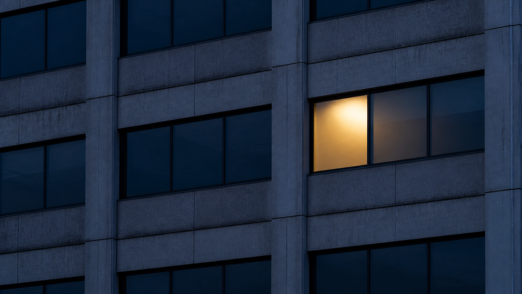 One empty frosted office window illuminated at blue hour among rows of dark windows.