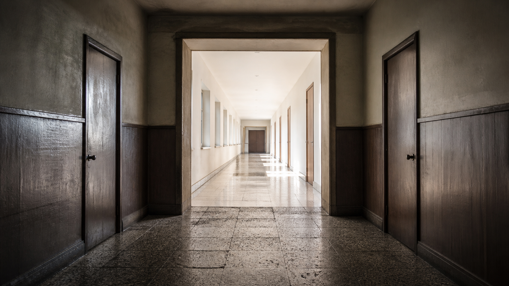 A dark institutional corridor opens through a broad threshold into a bright hallway.
