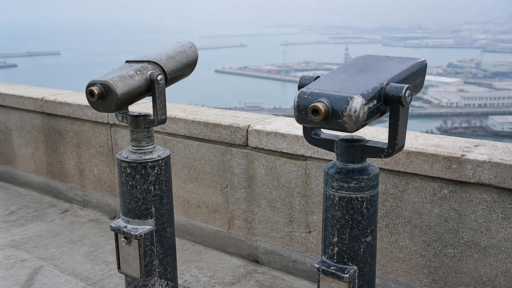 Two weathered public observation telescopes with different bodies point toward the same distant harbor.