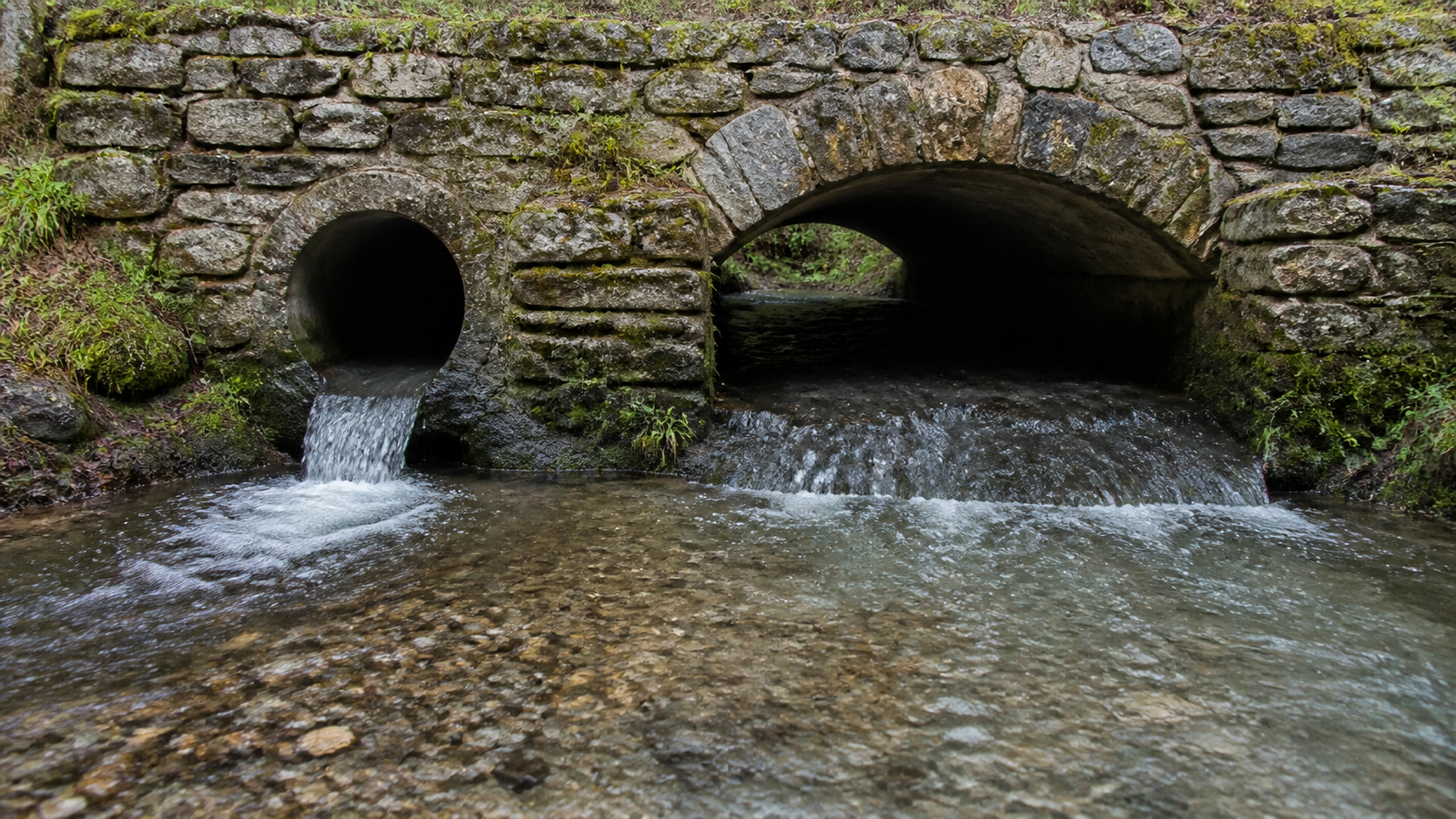 Clear water leaves one narrow round culvert and one broad stone arch into the same gravel stream.