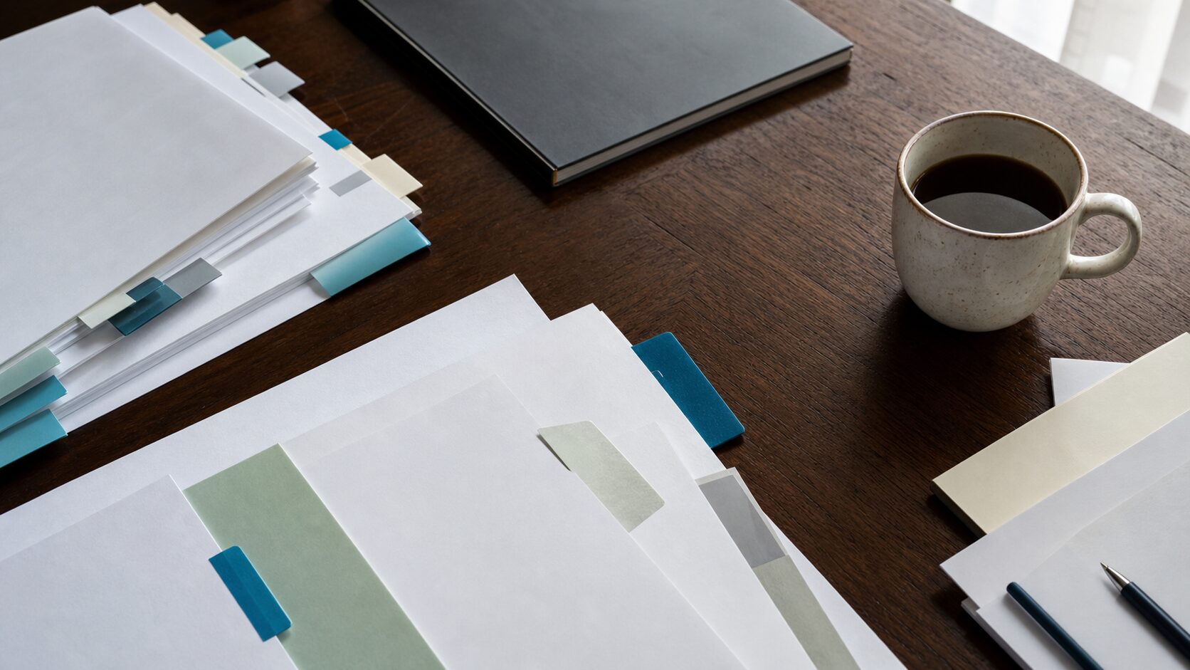Blank research papers with muted tabs, a closed notebook and a coffee cup on a dark wooden table