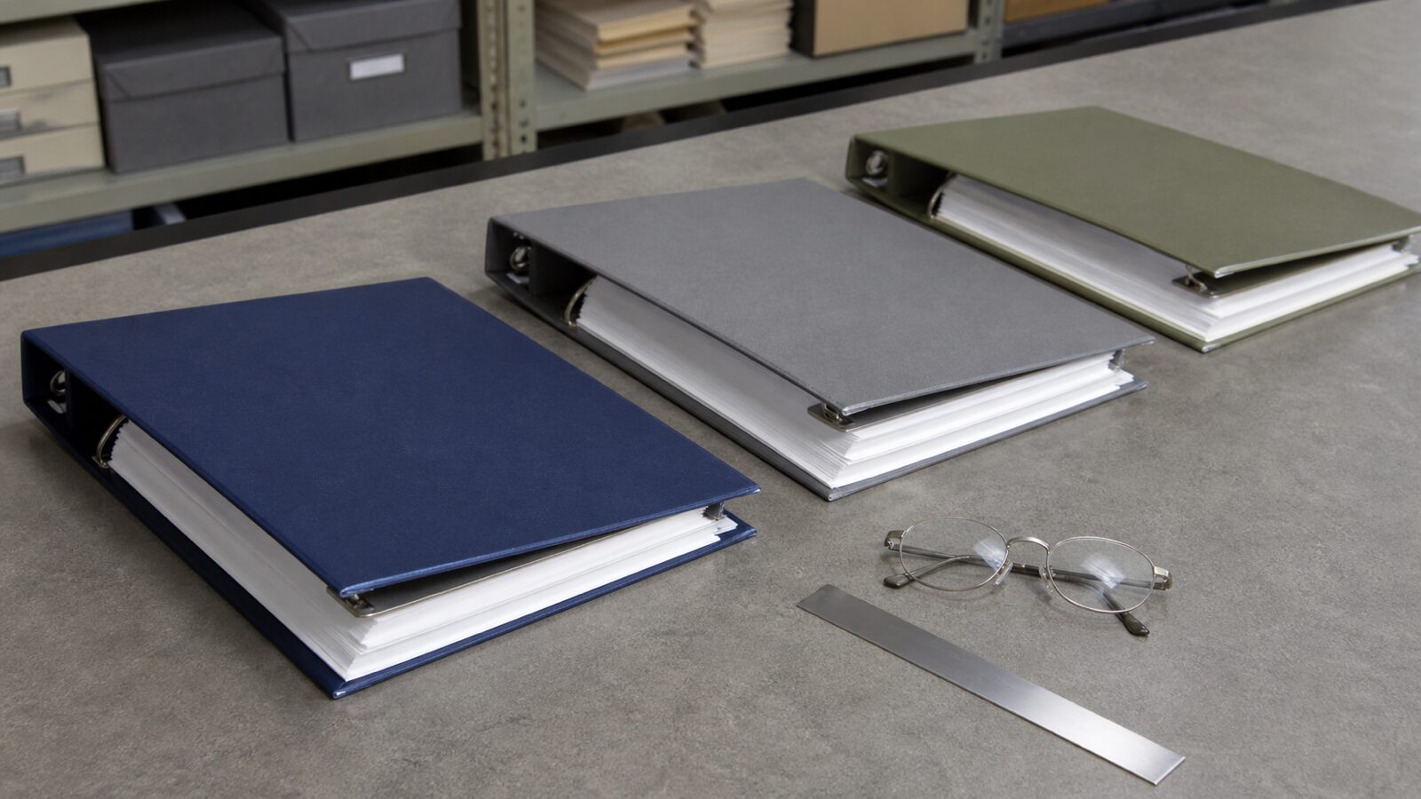 Three plain binders, reading glasses and a metal ruler arranged on an archive-room workbench