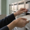 Hands place a blank envelope into an unbranded brushed-metal mailbox in a quiet mailroom.