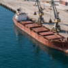 An unbranded cargo vessel lies beside a concrete quay and loading cranes in a working harbor.