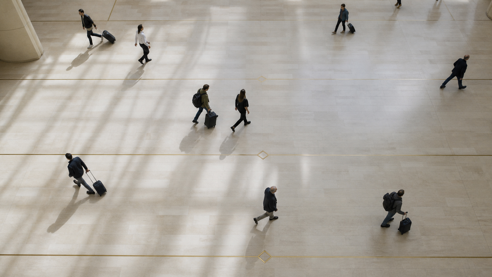 Travelers with small rolling luggage cross a sunlit limestone concourse seen from above