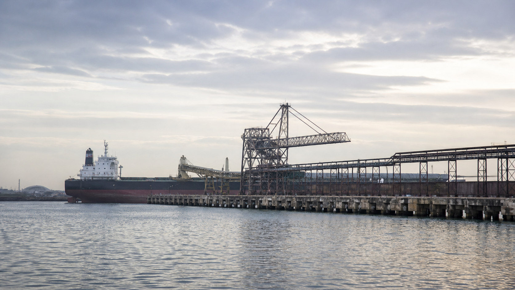 Unbranded dry-bulk ship beside a conveyor pier viewed across calm harbor water