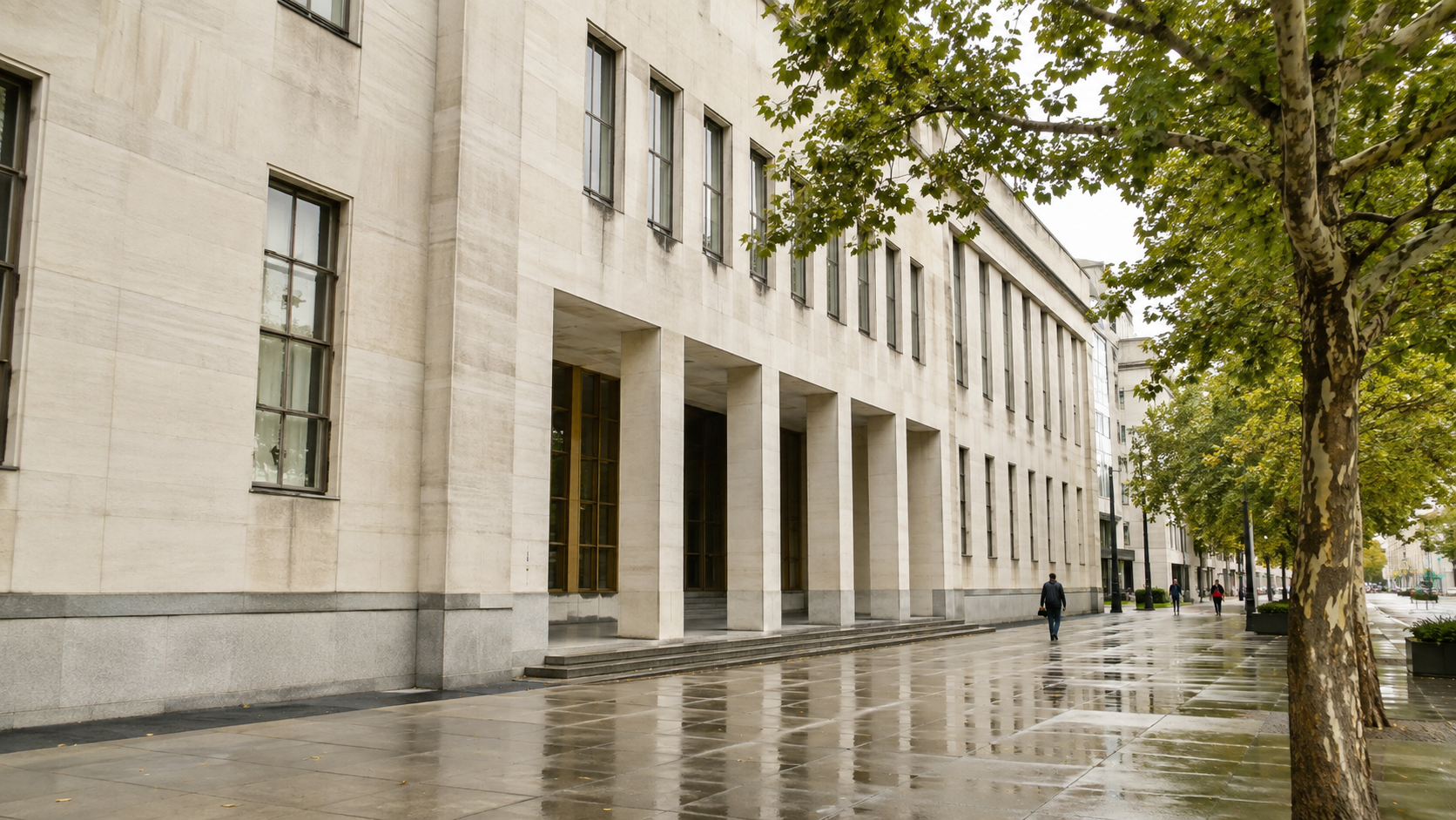 An unbranded pale-stone civic building beside a rain-damp tree-lined sidewalk