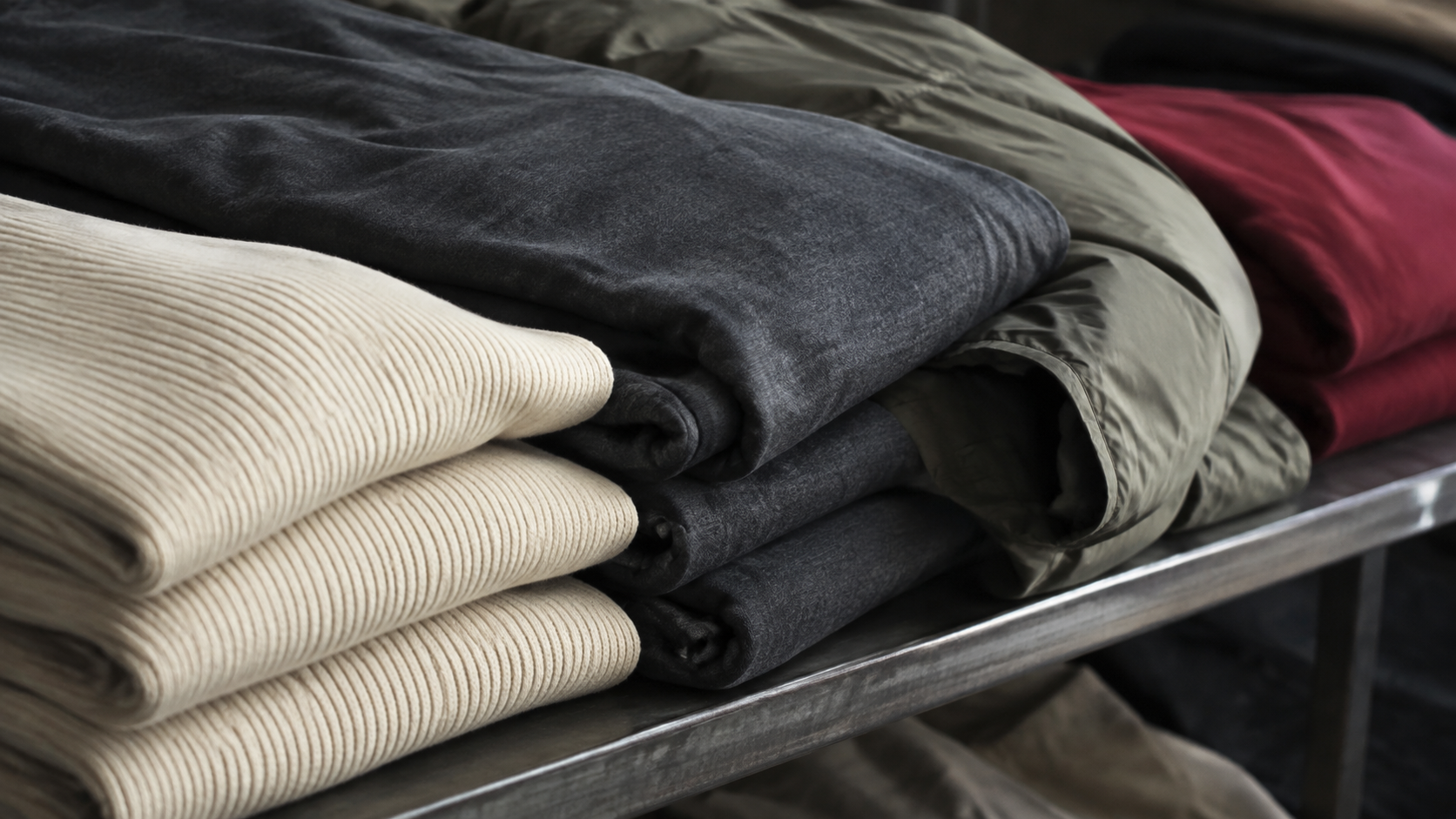 Close view of unbranded cream, charcoal, olive and deep-red folded apparel fabrics on a metal retail shelf