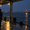 A commuter standing under a rain-wet covered pier at blue hour, facing dark harbor water and a distant terminal light.